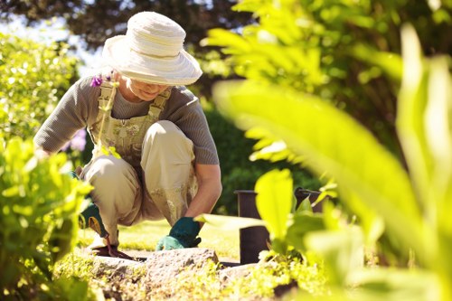 A gardener wearing a wide-brimmed white hat, glasses, and gardening overalls is kneeling on a well-maintained lawn in a landscaped outdoor space, engaging in garden maintenance. The scene shows the lower half of the gardener, who is using gardening tools on soil beds bordered by bricks. Surrounding the area are lush green plants, bushes, and trees that provide vibrant foliage and natural shade. The background features a neatly trimmed hedge and a variety of flowering plants, contributing to the aesthetic appeal of the garden. The paving stones in the foreground are textured and clean, and the overall scene suggests a bright, sunny day with natural light highlighting the different shades of green and earthy tones. This outdoor environment appears to be part of a private garden, possibly in the Ealing area, maintained by a professional gardening service like Hedge Trimming Ealing, emphasizing the importance of garden upkeep and landscaping quality.